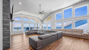 A spacious living room with a large L-shaped sectional sofa, a ceiling fan, and large windows offering ocean views. - Premier Soundfront w/ Pool, Hot Tub, Elevator, EV Charger, Theater & Game Rooms (Avon)