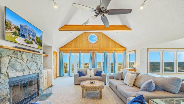 A spacious living room with a stone fireplace, a ceiling fan, and large windows offering a view of the landscape. - Oceanfront Dog Friendly Home w/ Saltwater Pool, Elevator, Hot Tub, Cmty Perks (Waves)