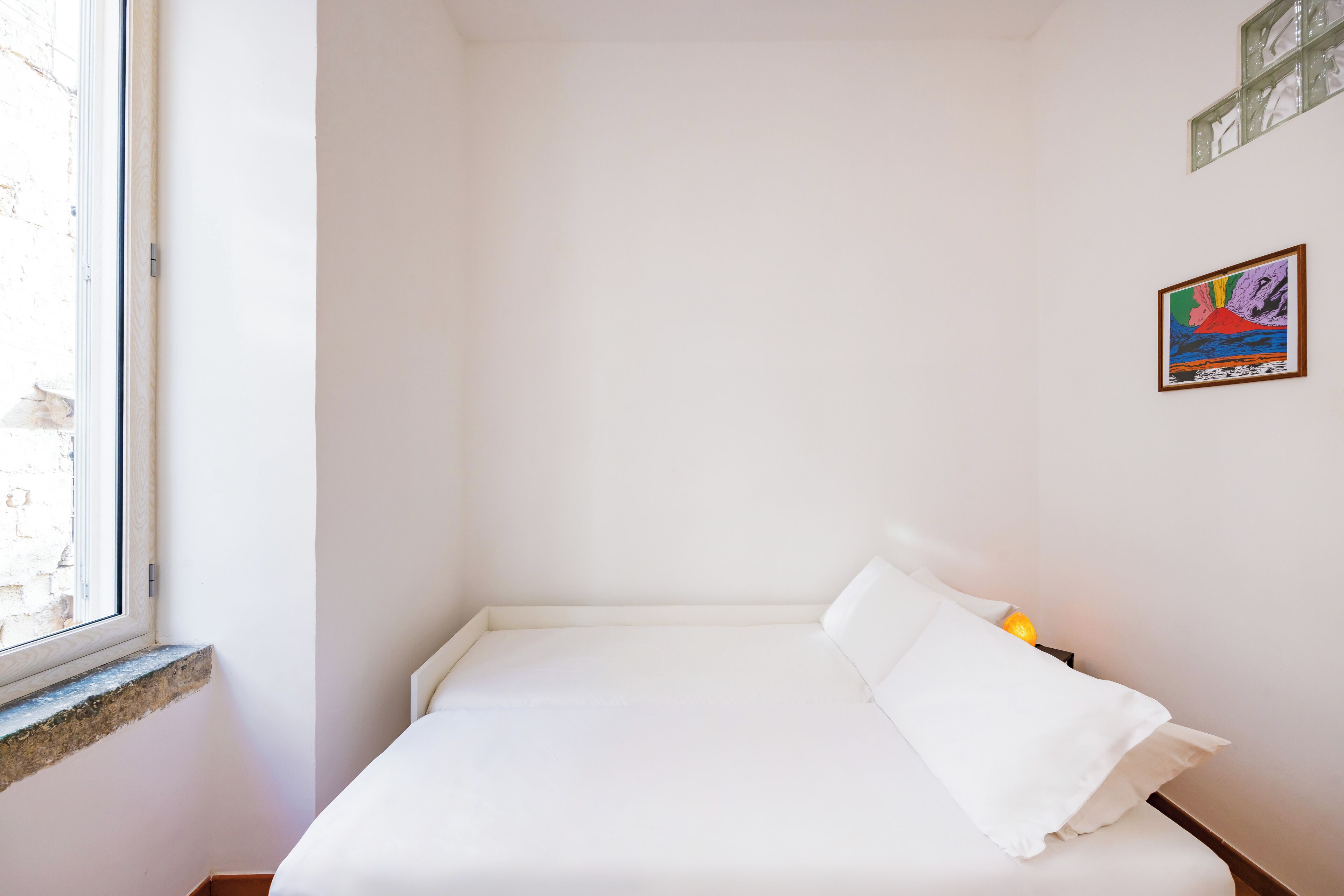 Quarto pequeno e branco, com uma cama de solteiro, janela, mesa de cabeceira e um quadro colorido na parede.