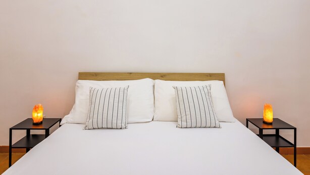 Ein Bett mit weißer Bettwäsche und gestreiftem Kissen, begleitet von zwei Nachttischen mit einer wärmelampe auf jedem.