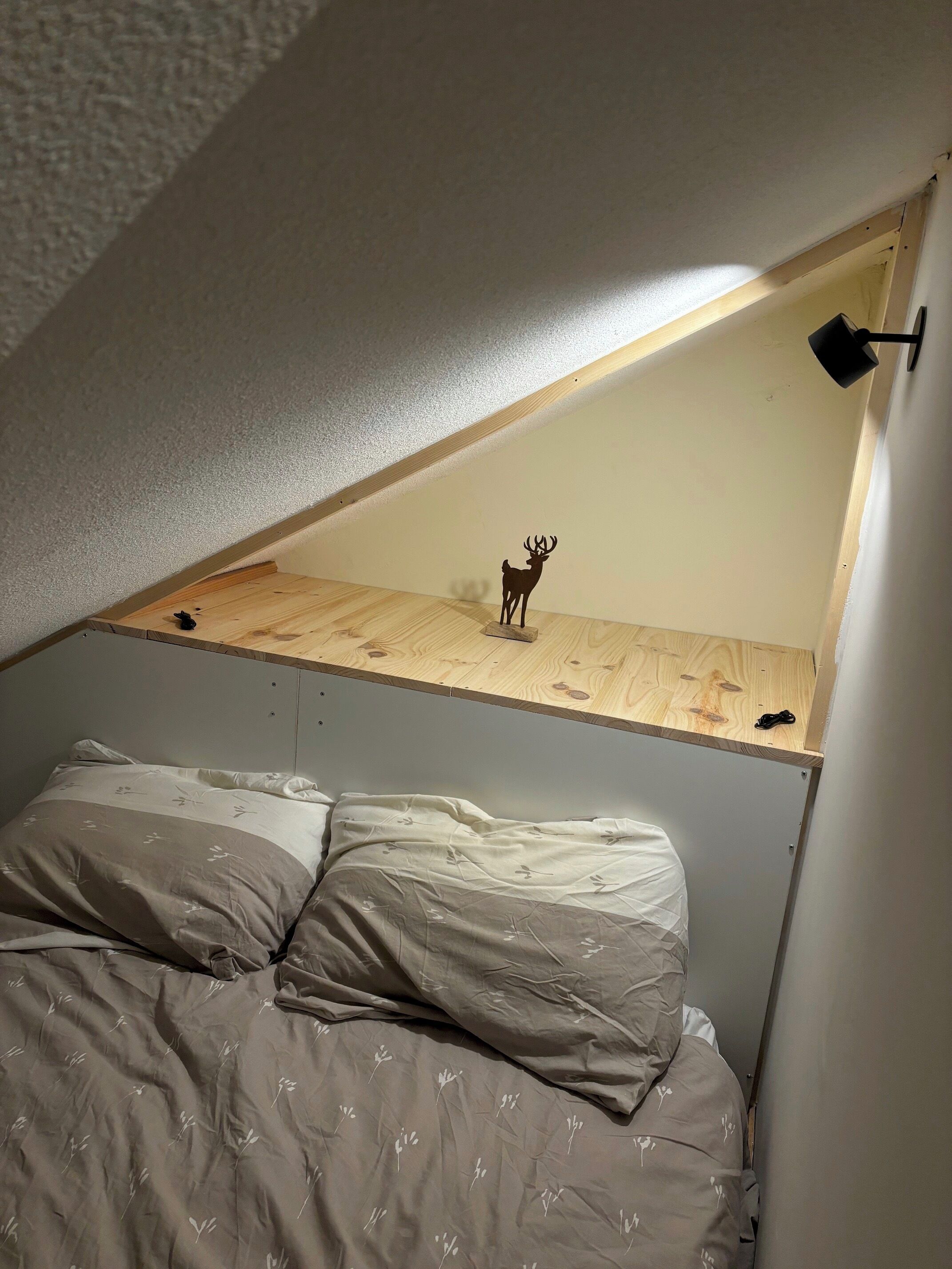 A bedroom with a sloped ceiling, a bed with grey floral bedding, a small wooden platform, a black lamp, and a deer figurine.
