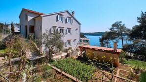 Une maison à deux étages avec un balcon donnant sur un lac, entourée de verdure et d’un jardin.