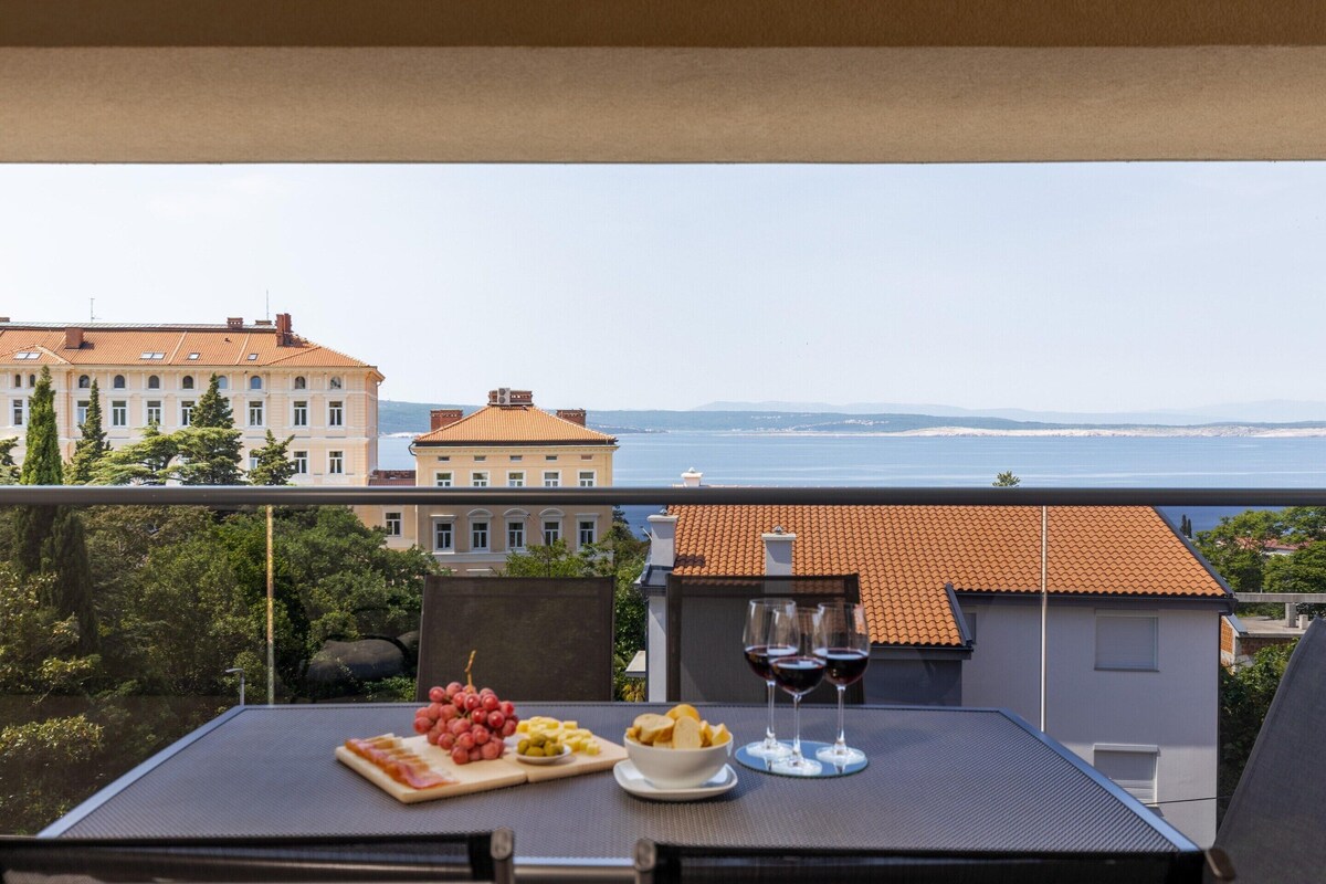 A balcony with a table set for a meal, including a plate of food and two glasses of red wine, overlooking a cityscape with buildings and a body of water.