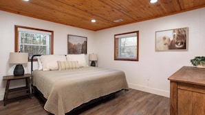 A bedroom with a wooden ceiling, a bed with a beige bedspread, two bedside tables with lamps, and two windows with blinds. - Walk Into The National Park/View (Sevierville)