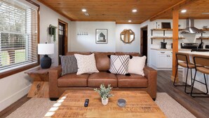 A cozy living room with a brown leather sofa, a wooden coffee table, and a kitchen area with white cabinets and a stainless steel range hood. - Walk Into The National Park/View (Sevierville)