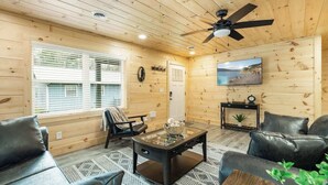 A cozy room with wooden walls, a ceiling fan, a large window with blinds, a sofa, a coffee table, and a TV mounted on the wall.