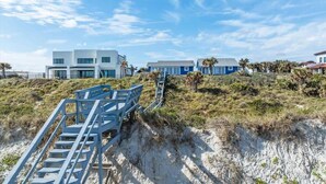 Et kystnært bygningskompleks i moderne arkitektur, en trappe ned til en strand, og en klar himmel.