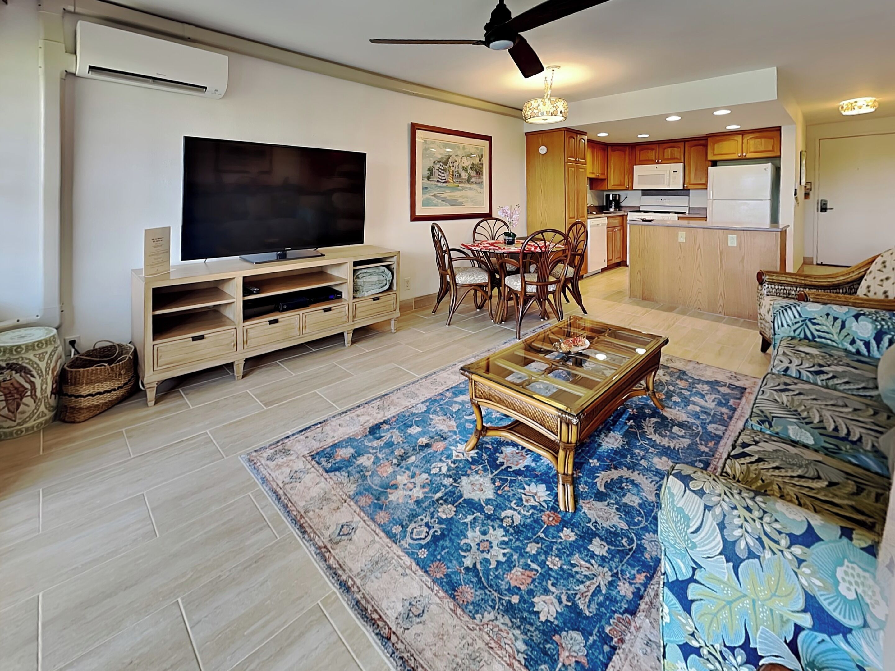 Sala de estar con televisor de pantalla plana, mueble de entretenimiento de madera, mesa de comedor con sillas, sofá estampado y área de cocina con gabinetes de madera y ventilador de techo.