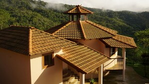 Un bâtiment traditionnel, doté d’un toit en tuiles et surmonté d’une petite structure en forme de tour, le tout entouré de collines verdoyantes.
