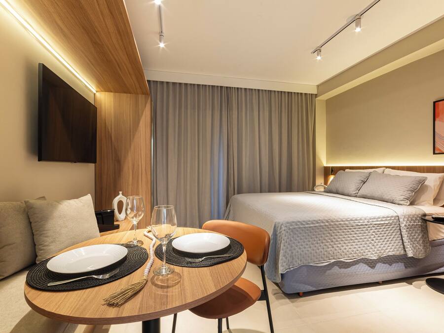 A modern hotel room with a neatly made bed, a dining table with plates and cutlery, and a television mounted on the wall.