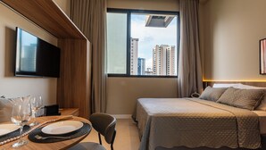 A hotel room with a bed, a TV, a dining table with plates and glasses, and a view of buildings through the window. - Lexy Hotel (São Paulo)