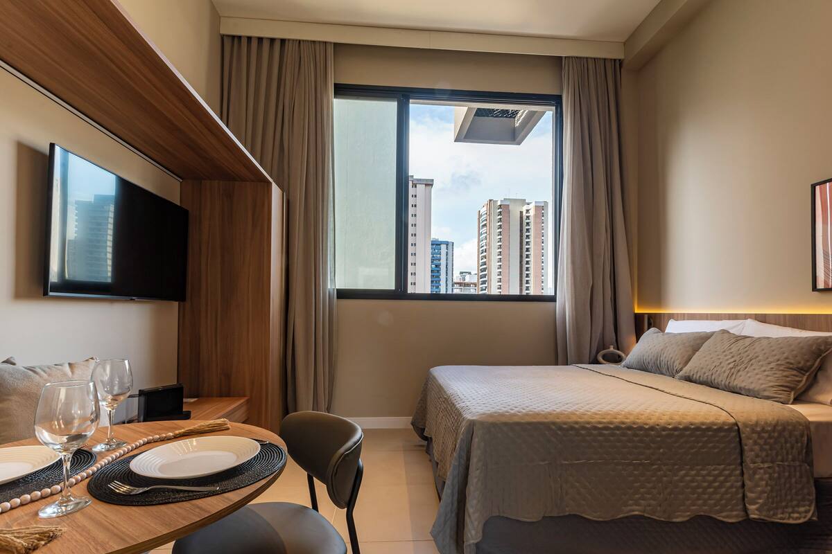A hotel room with a bed, a TV, a dining table with plates and glasses, and a view of buildings through the window.