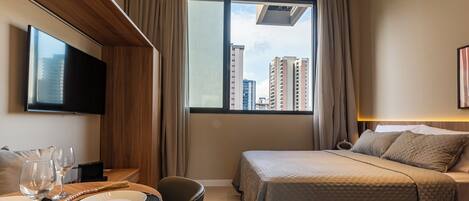 A hotel room with a bed, a TV, a dining table with plates and glasses, and a view of buildings through the window.