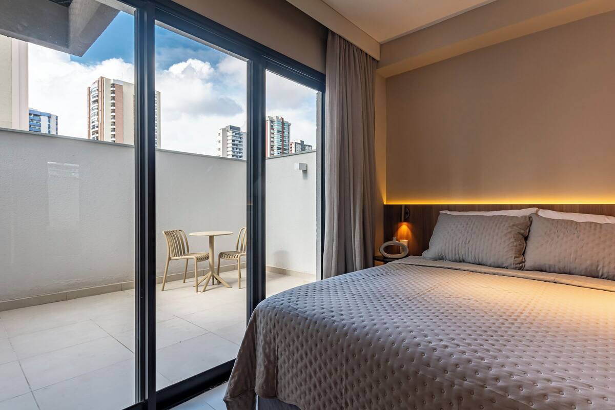 A modern hotel room with a large bed, a small table, and chairs, a balcony with a view of the cityscape.
