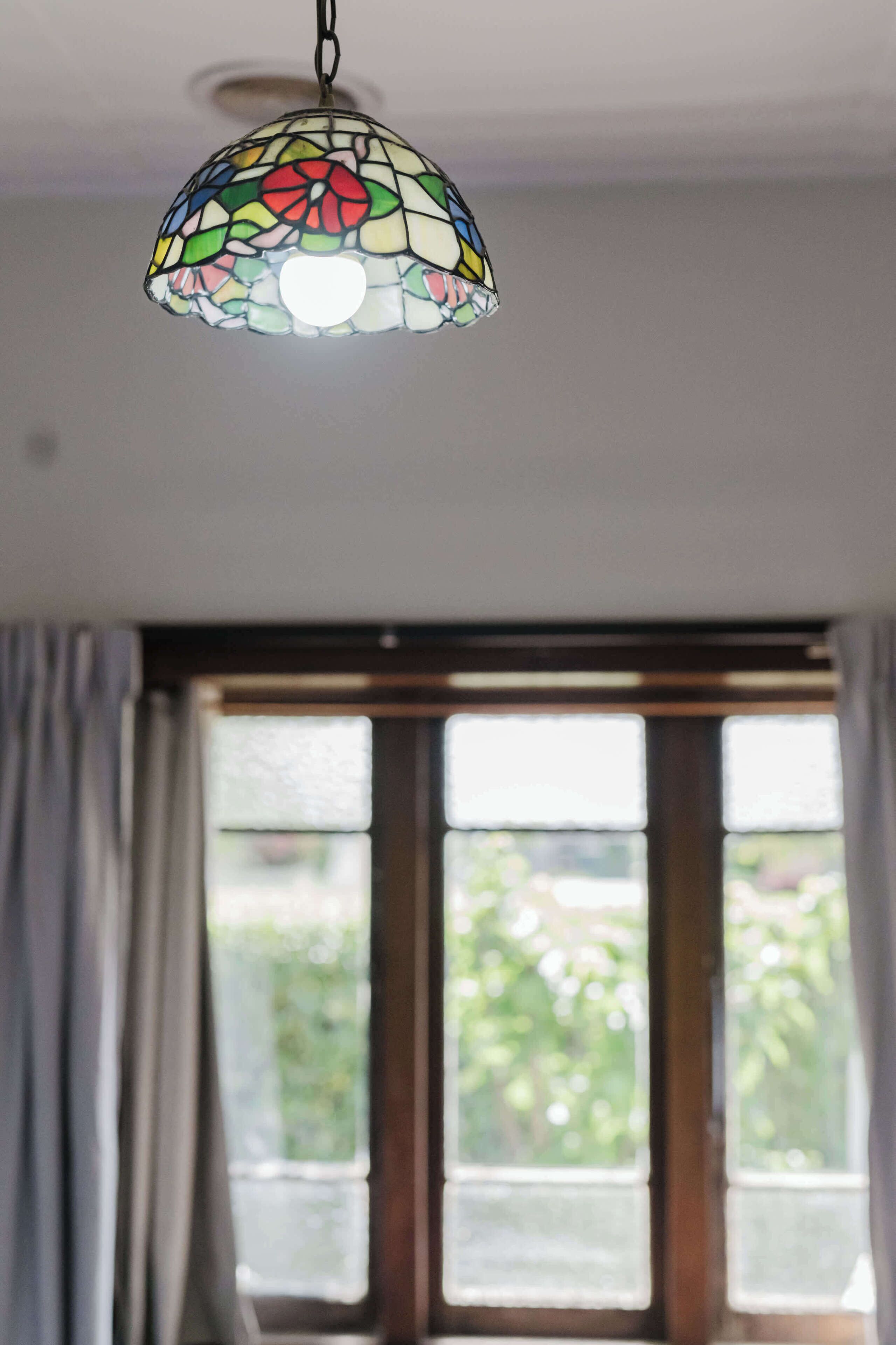 Ein Zimmer mit großem Fenster, einer hängenden Leuchte aus Buntglas und einem Vorhang.