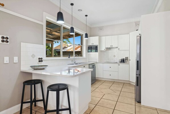 Une cuisine moderne avec des meubles blancs, un grand îlot central doté de tabourets, un éclairage suspendu et une vue sur l’extérieur.