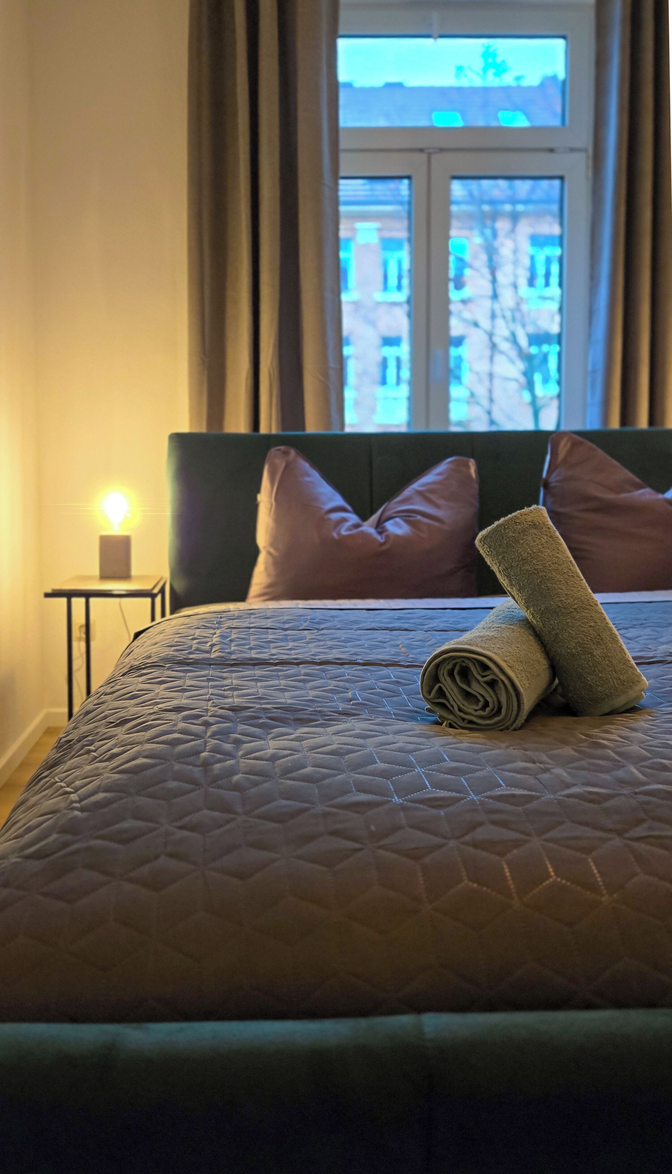 Una cama bien hecha con cubrecolcha acolchado, dos almohadas y una toalla enrollada en una habitación de hotel.