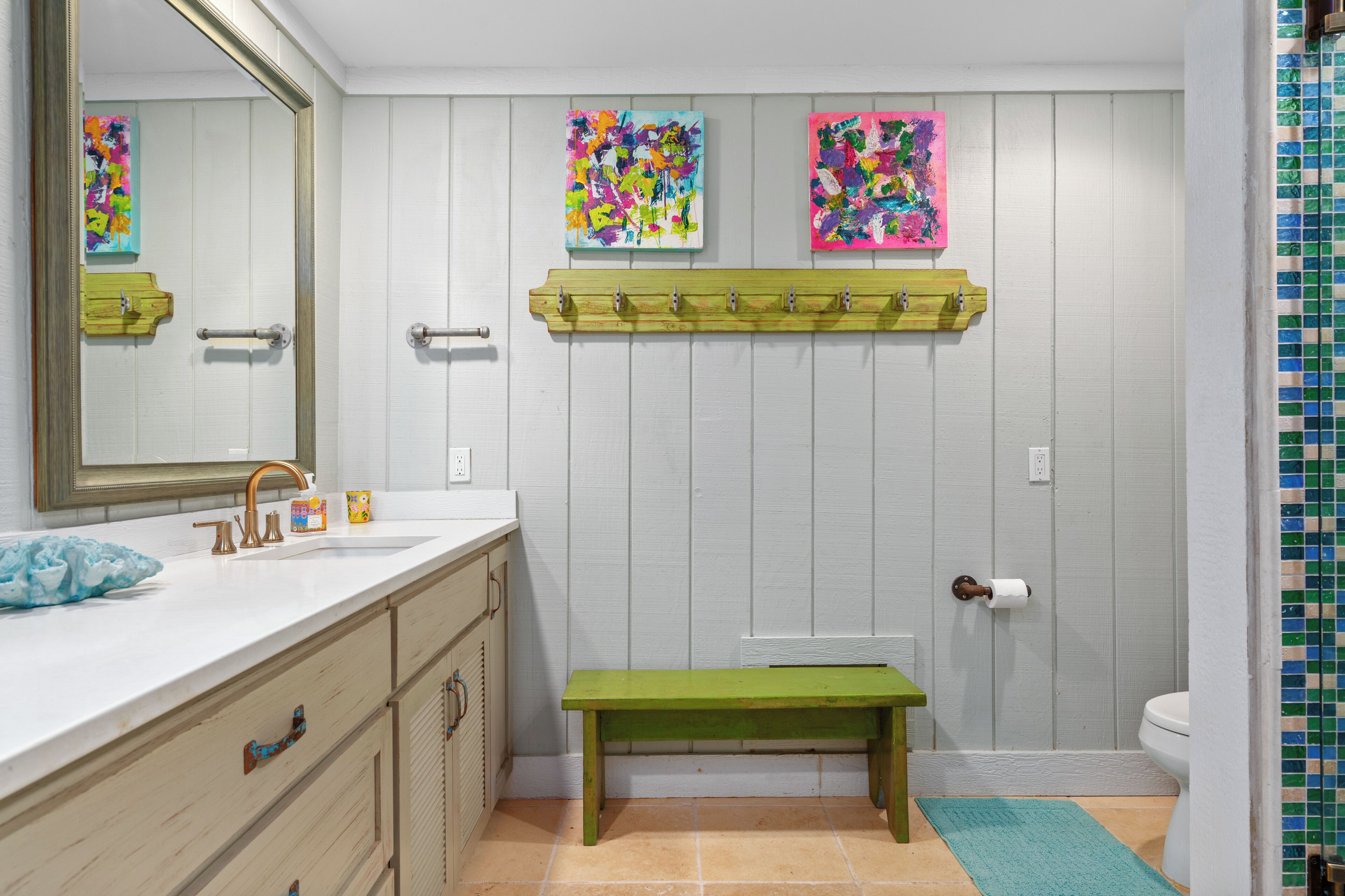 Un baño con tocador blanco, un banco verde y dos cuadros abstractos y coloridos en la pared.