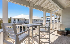 A covered outdoor area with white wooden furniture, including chairs and a table, overlooking a row of white buildings with balconies.