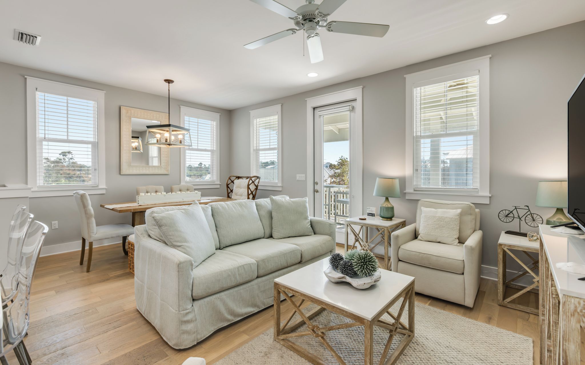 A spacious living room with a large sofa, a coffee table, and a television. There are multiple windows providing natural light and a view of the outdoors.