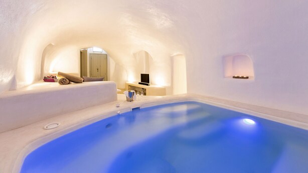 A modern, minimalist room with a large blue pool, a cozy seating area, and a television. - Kampanario Luxury Villa (Thira - Santorini)
