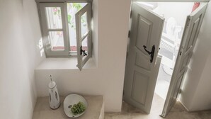 A room with a white door, a window with a view of pink flowers, a white plate with green grapes, and a white object on the floor. - Kampanario Luxury Villa (Thira - Santorini)