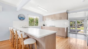 A modern kitchen with a central island, wooden chairs, and a view of the outdoors. - A Touch Of Paradise - 4 Min Walk to Beach (Callala Beach)