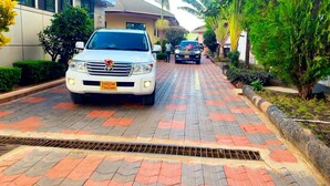 A paved driveway leading to a building with a white car parked in front. - Lenny Hotel Ltd. (Geita)