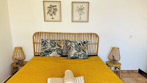 Una cama con una colcha amarilla con textura, dos almohadas con estampado de hojas tropicales y una toalla enrollada sobre la cama. Hay dos lámparas de pared a cada lado de la cama.