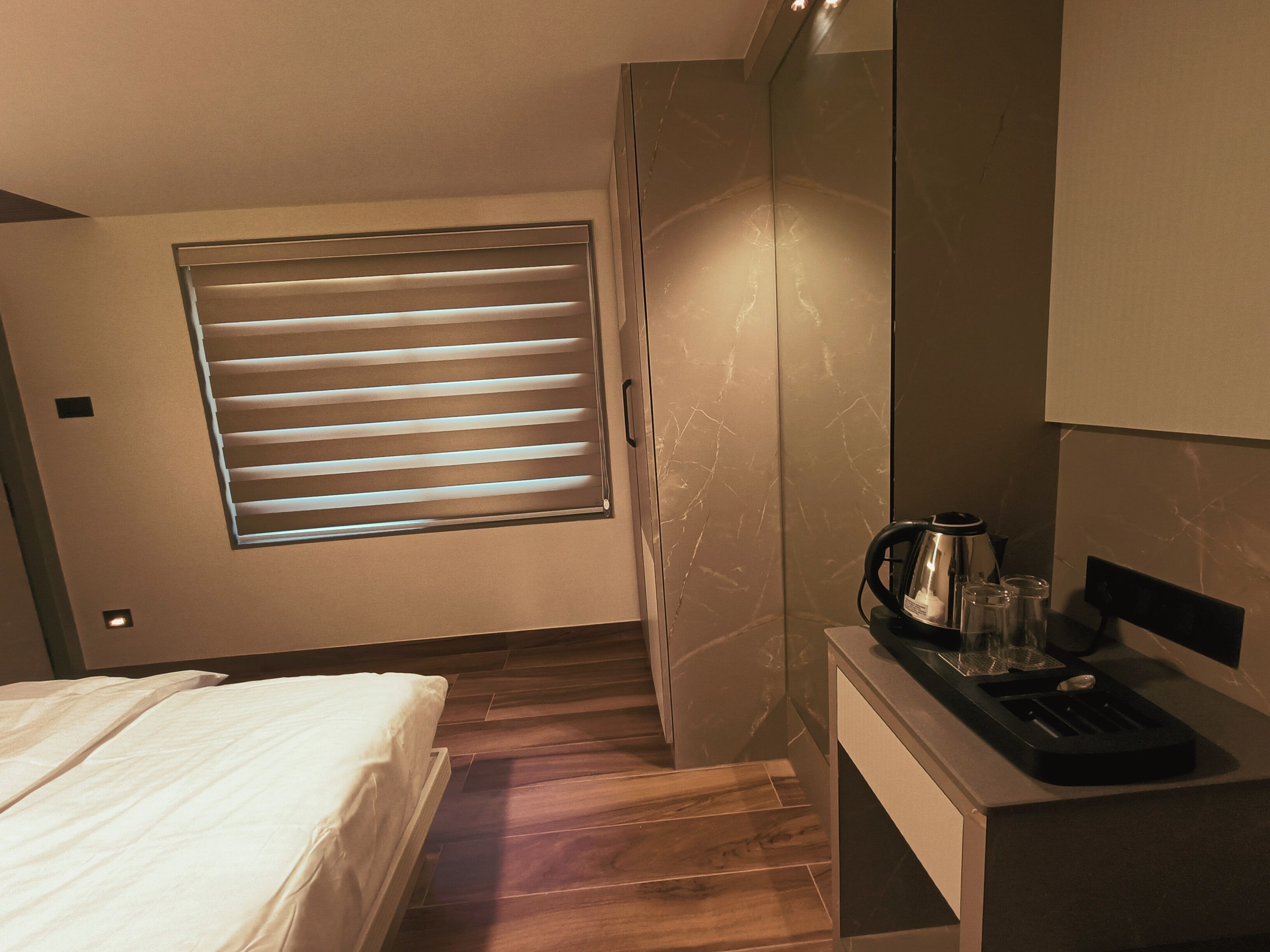 A hotel room with a bed, a small desk with a kettle and glasses, a window with blinds, and a marble wall.