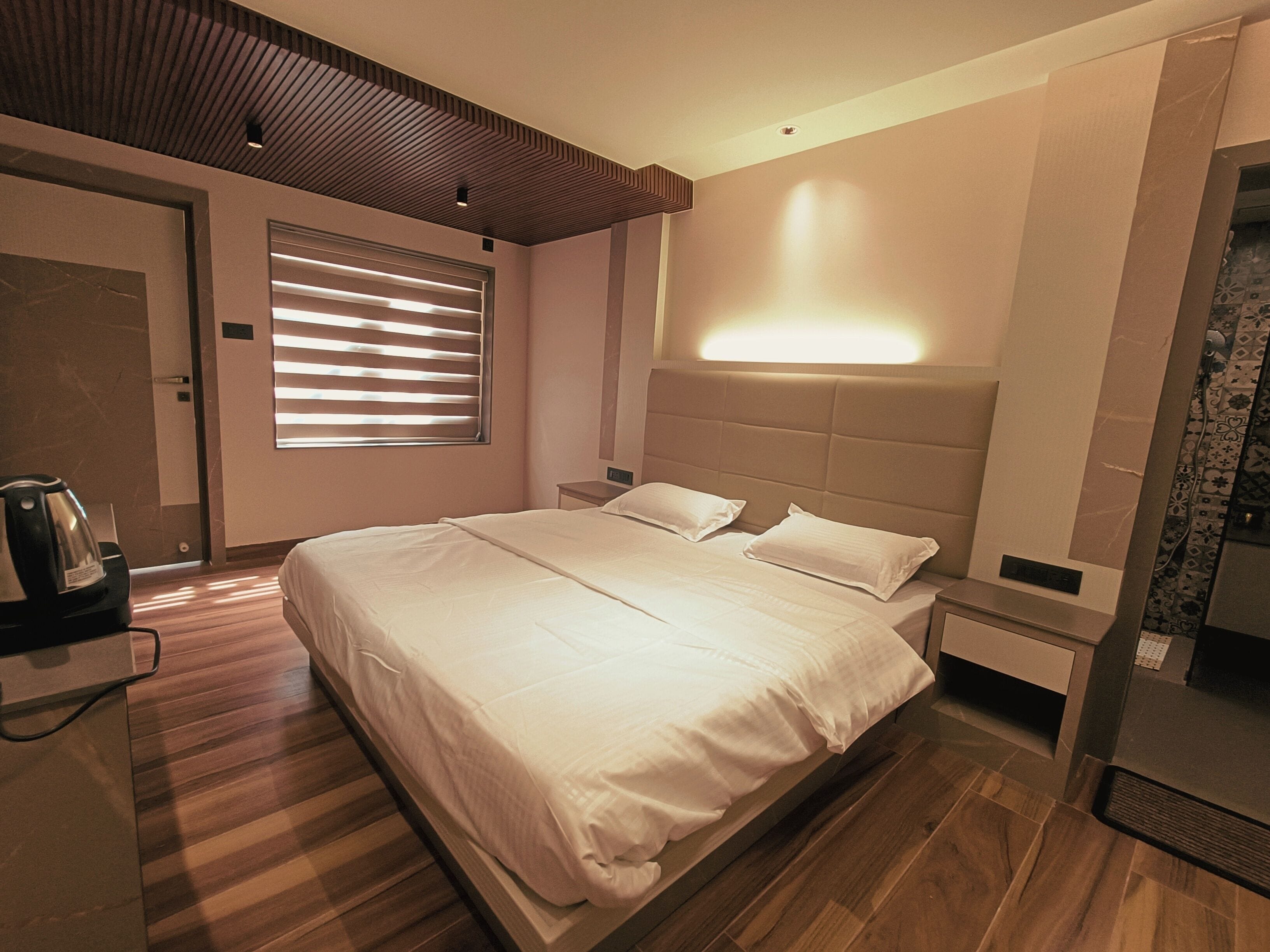 A modern hotel room with a large bed, bedside tables, a coffee maker, and a window with blinds.