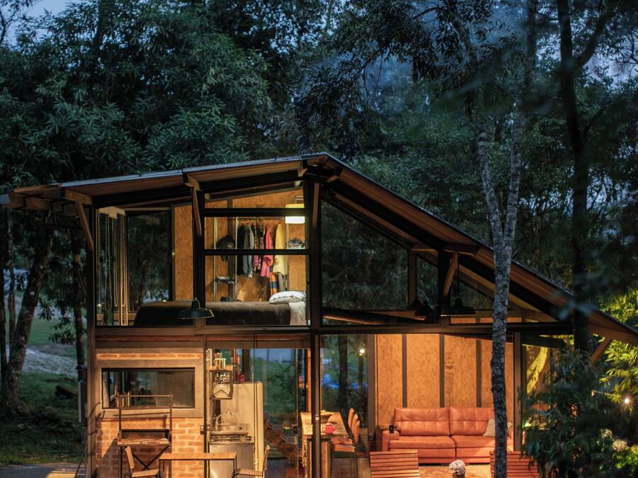 A modern wooden cabin with glass walls, a dining area, and a cozy seating space surrounded by lush greenery.