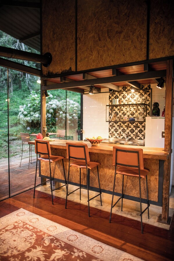 A modern kitchen with a bar area, wooden flooring, and a view of greenery outside. - Chale de Vidro nas montanhas (Itamonte)