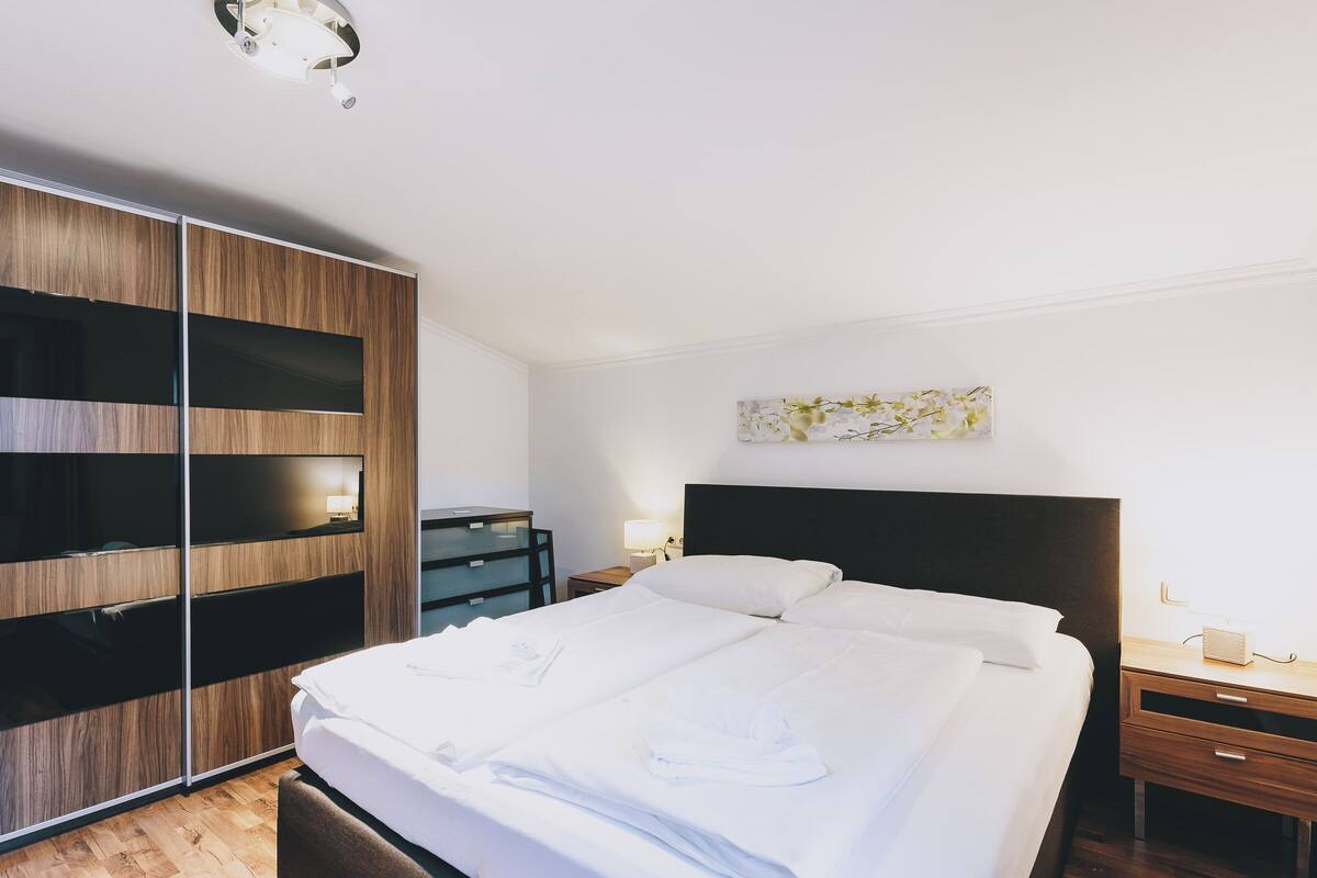 A modern hotel room with a large bed, a wardrobe with sliding doors, a bedside table with a lamp, and a wall-mounted artwork.
