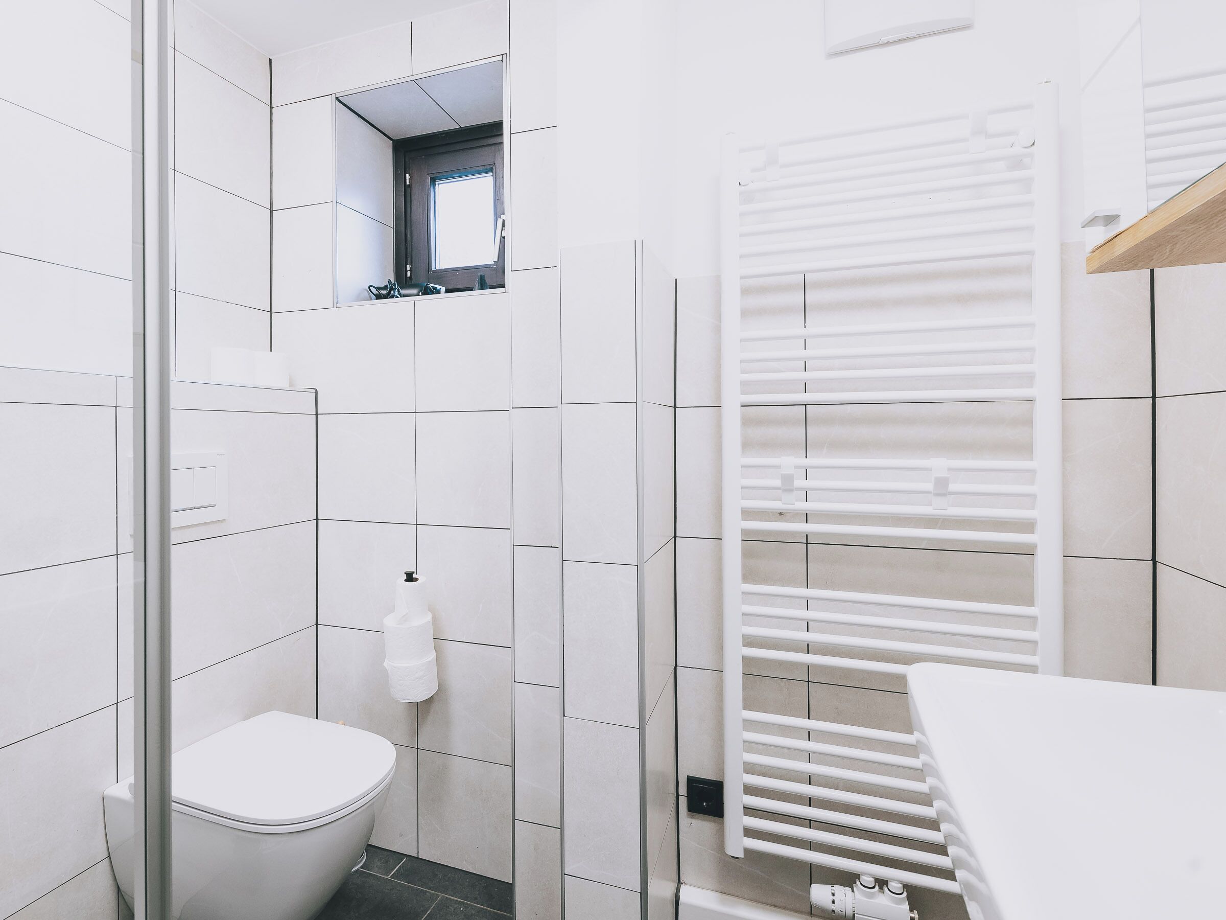 A modern bathroom with white tiles, a toilet, a glass shower enclosure, and a white towel rack.