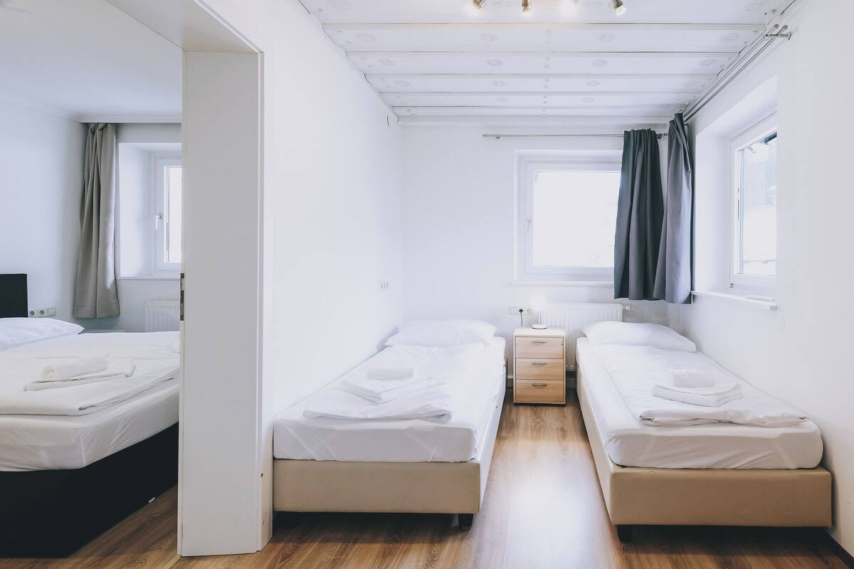 A hotel room with two single beds, a nightstand, a window with curtains, and a radiator.