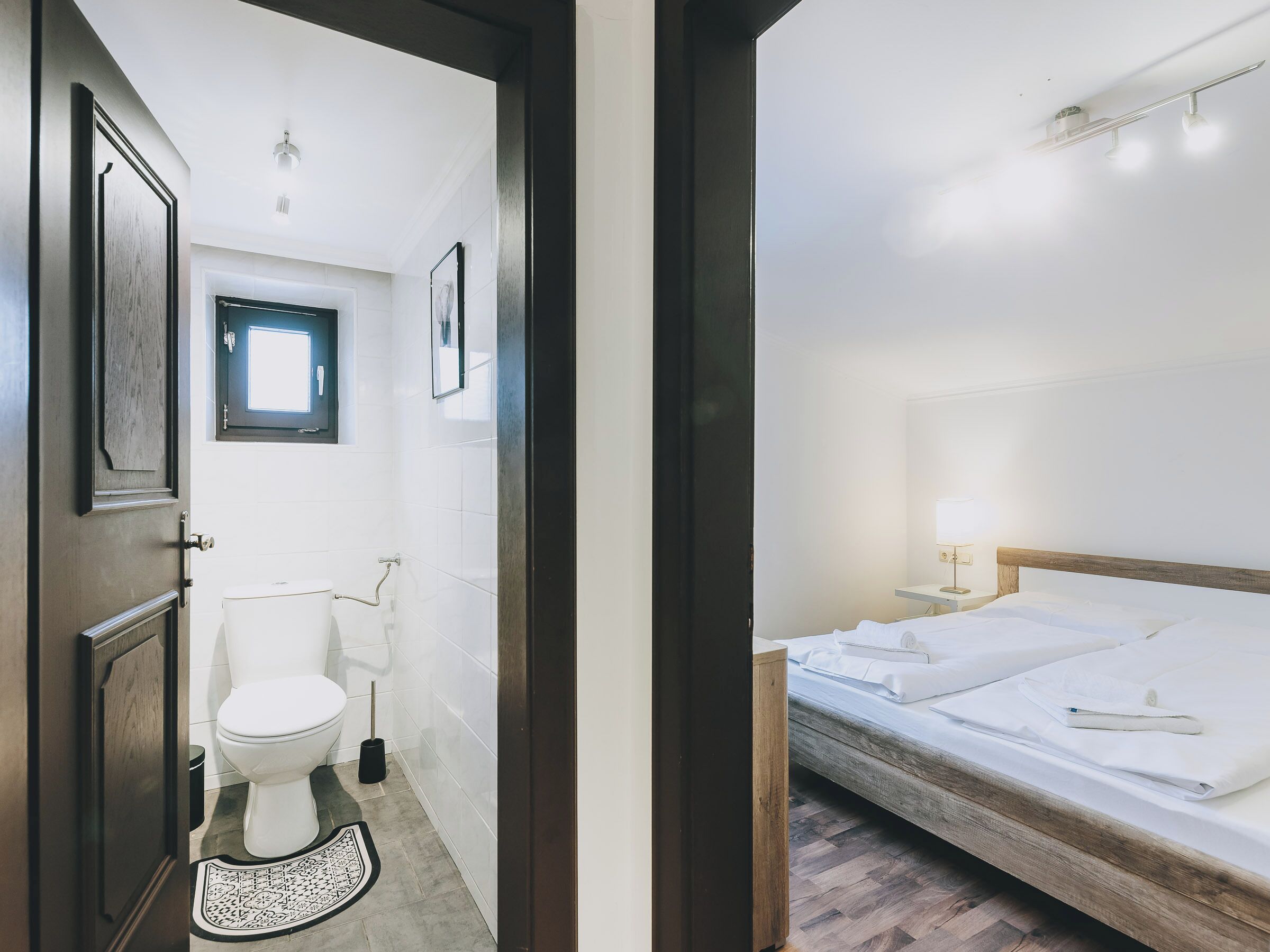 Une chambre d’hôtel avec un lit en bois, une table de chevet avec une lampe, une petite fenêtre et des toilettes équipées d’un bidet.