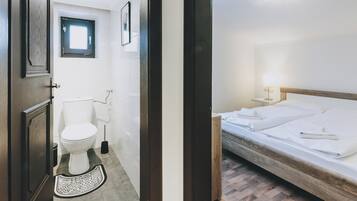 Une chambre d’hôtel avec un lit en bois, une table de chevet avec une lampe, une petite fenêtre et des toilettes équipées d’un bidet.