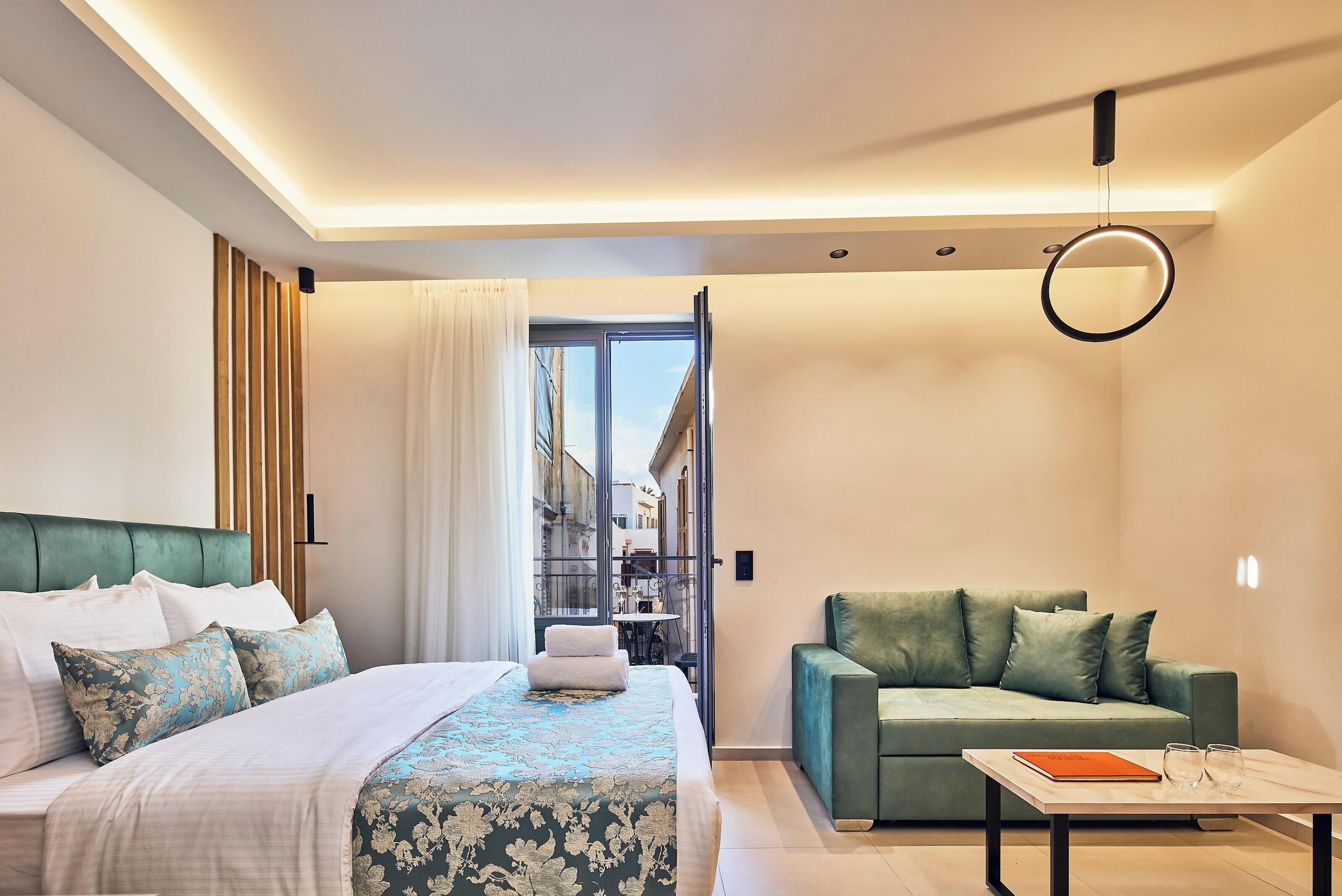 A modern hotel room with a bed, a green sofa, a small table, and a view through a glass door.