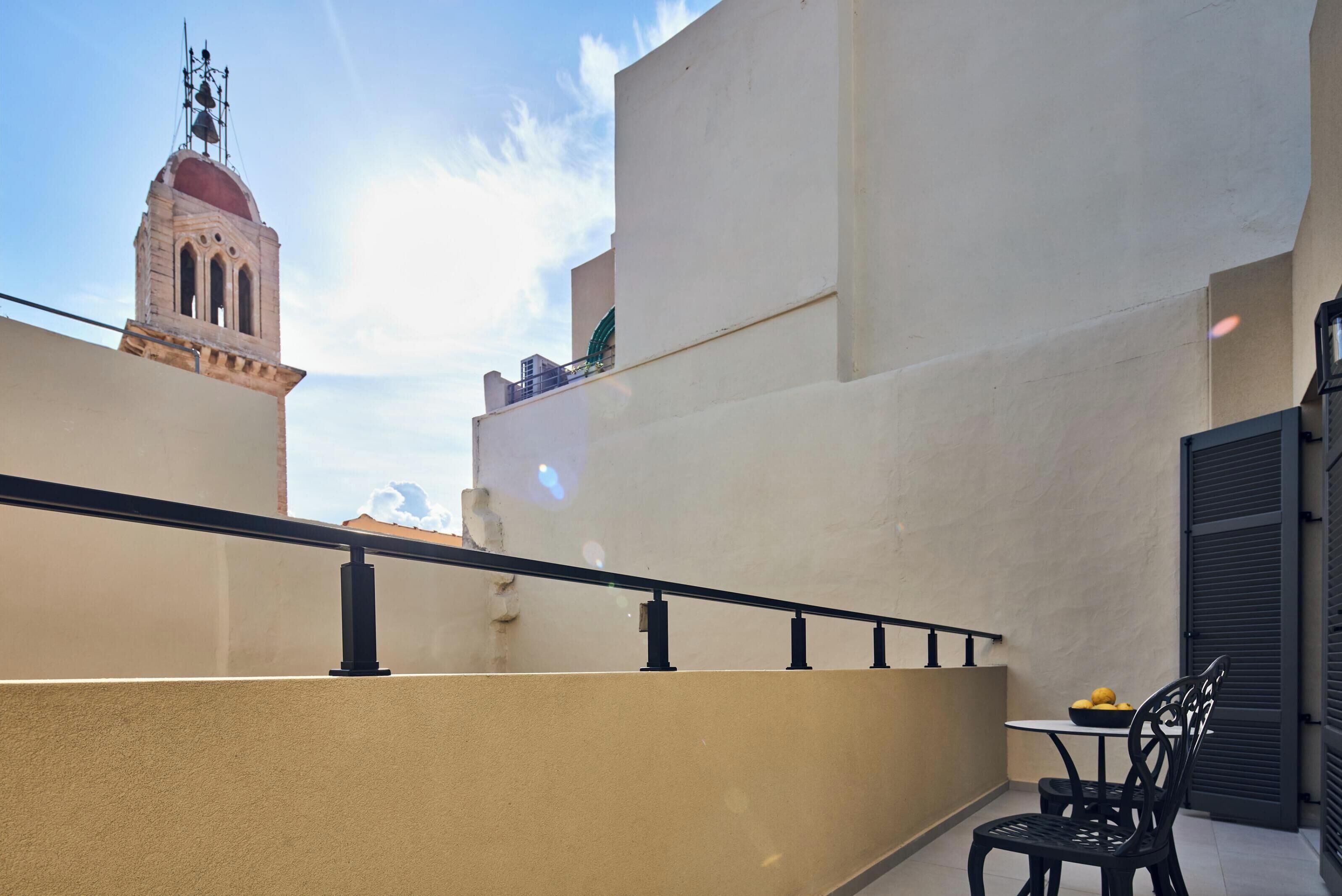 Un balcón con barandilla, una mesa con sillas y vistas a un edificio con una torre campanario.