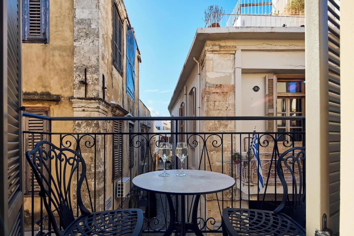 Une ruelle étroite avec une table ronde et deux chaises, bordée de vieux bâtiments dotés de balcons et de volets.