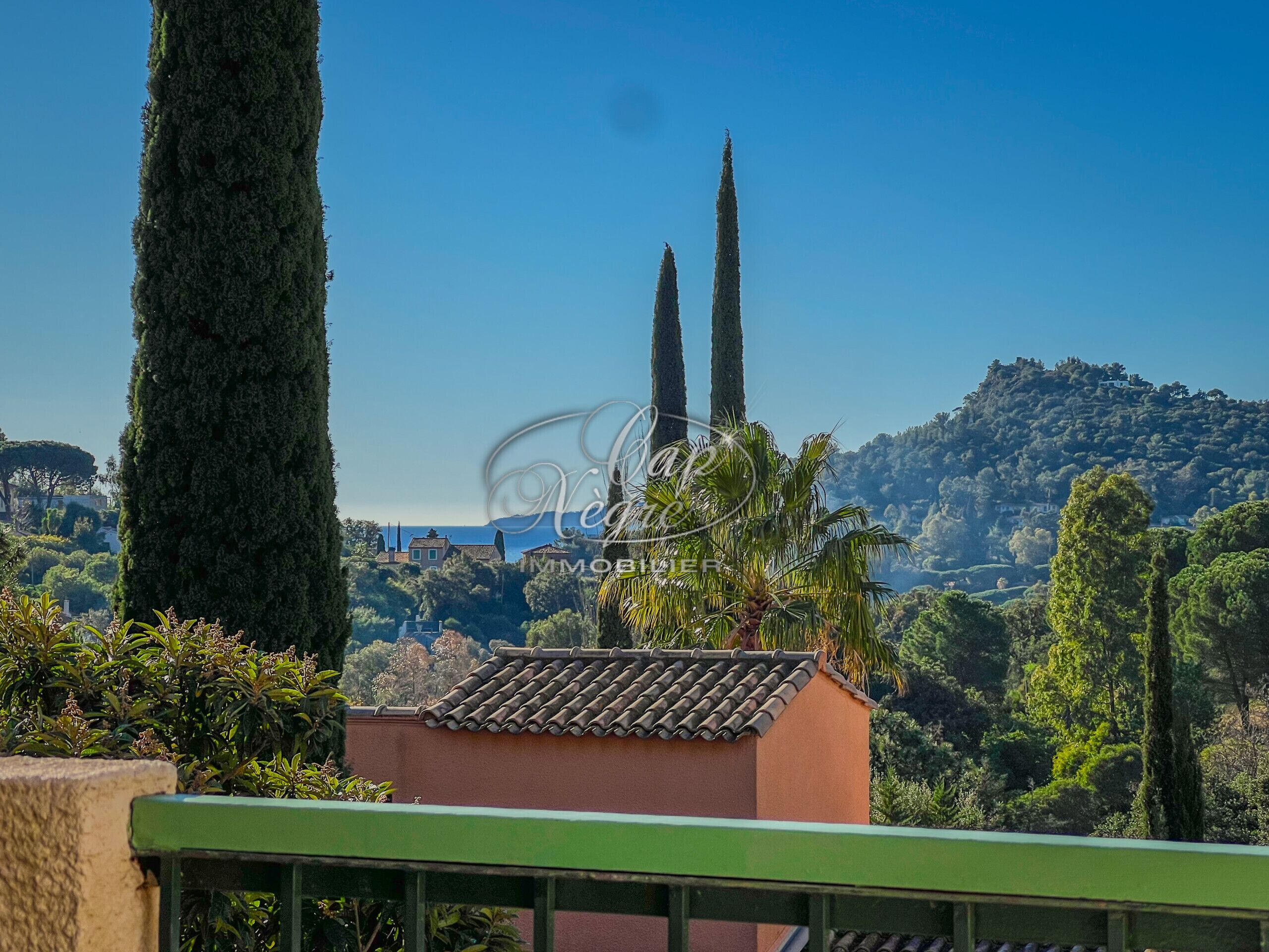 Une vue d’une villa avec un toit en tuiles, entourée d’une végétation luxuriante et de grands cyprès.