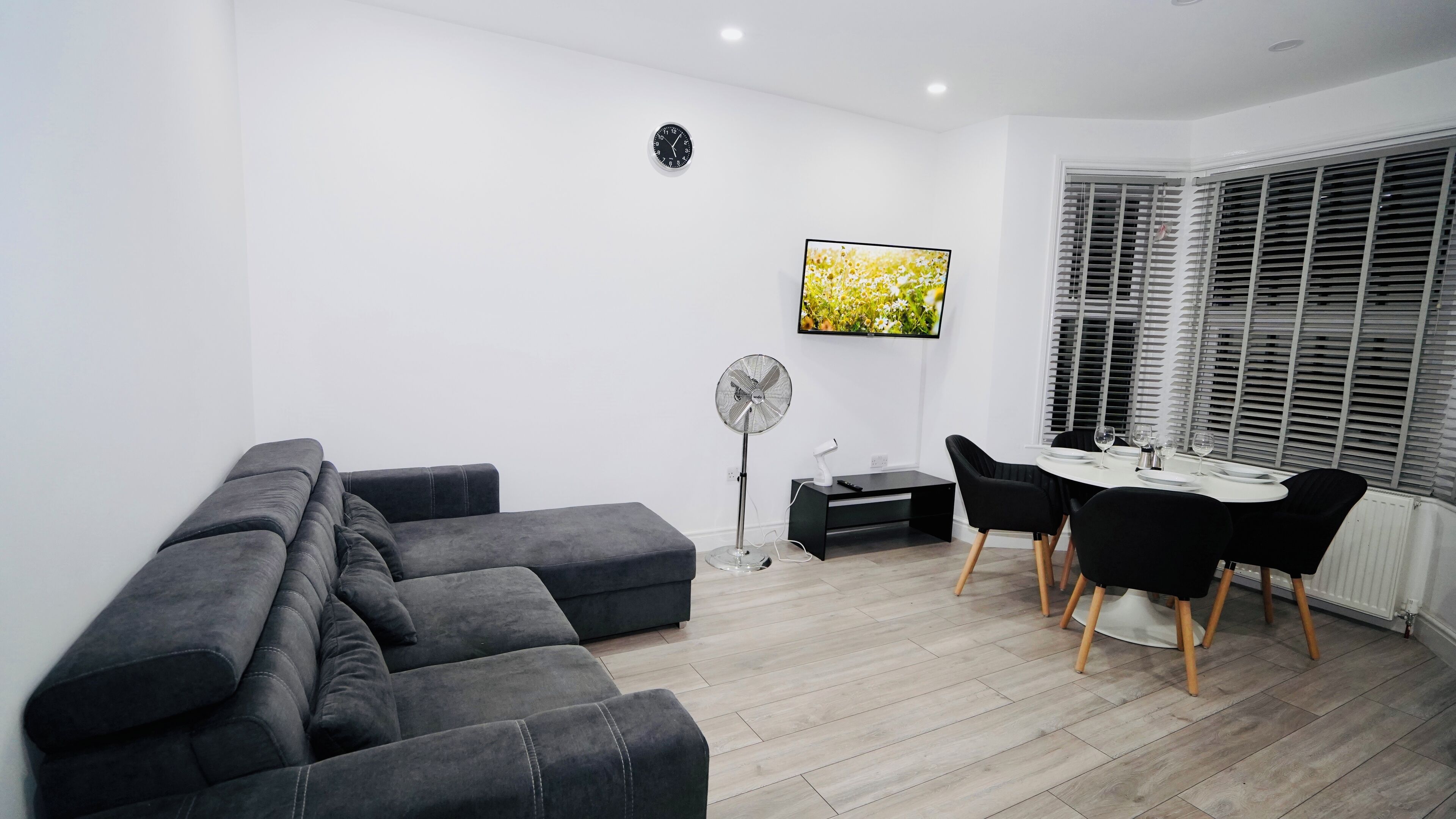 A modern living room with a grey sectional sofa, a dining table with four chairs, a standing fan, and a flat-screen TV mounted on the wall.