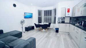 A modern kitchen with a built-in oven and a coffee machine, a dining area with a round table and black chairs, and a living area with a grey sofa and a floor fan. - ILford Station (Ilford)