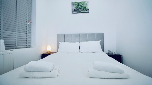 A neatly made bed with white linens and towels, a grey upholstered headboard, a bedside table with a lamp, and a small picture on the wall. - ILford Station (Ilford)