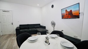A modern living room with a black sofa, a dining table set with plates and wine glasses, a wall-mounted fan, and a large painting of a desert landscape. - ILford Station (Ilford)