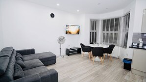 A modern living room with a sofa, a dining table with chairs, a TV, and a window with blinds. - ILford Station (Ilford)