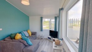 A modern living room with a grey sofa, a flat-screen TV, and a large window with curtains. - D37 Irene, Riviere Towans (Hayle)