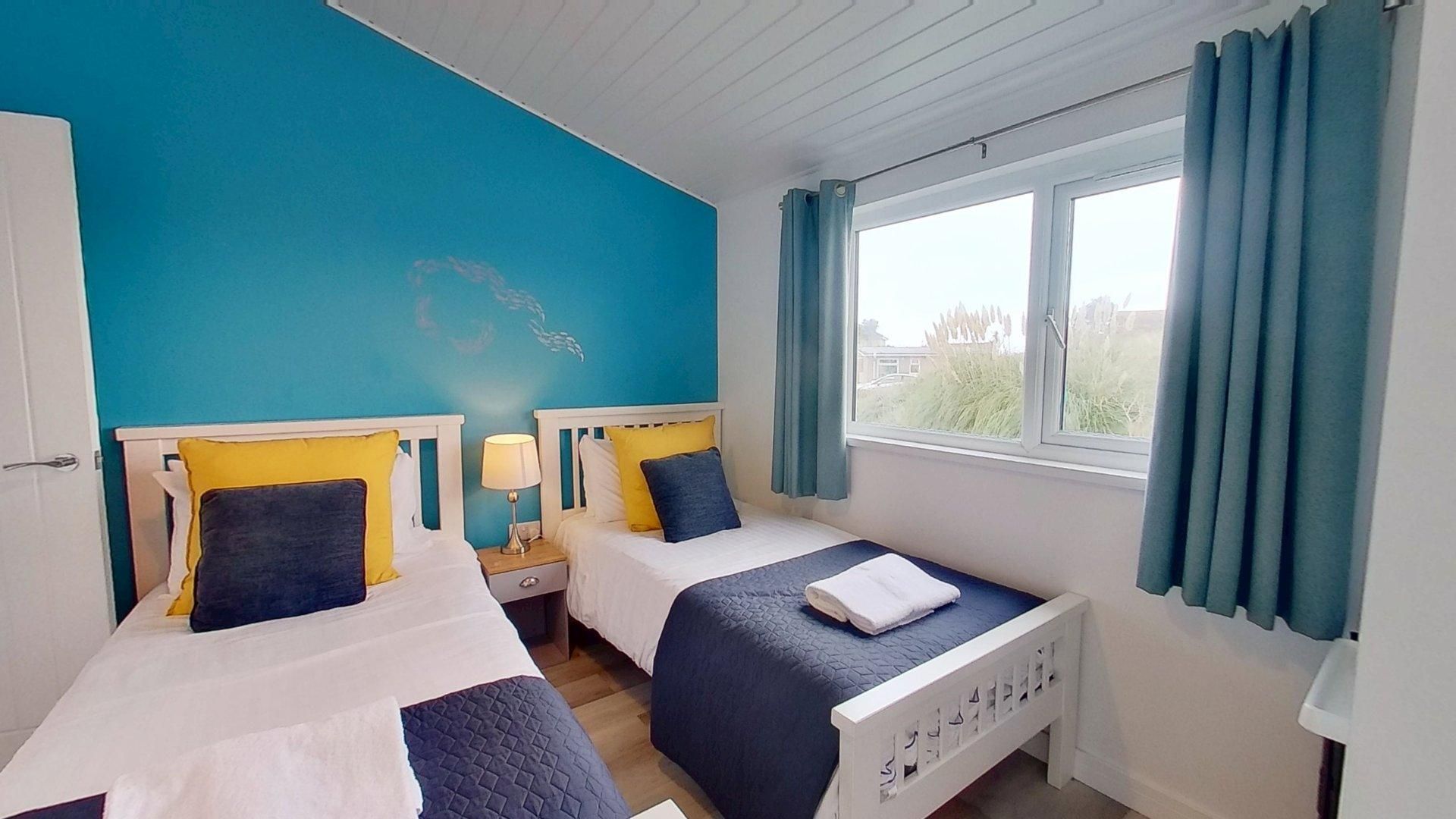 A room with two beds, a window with curtains, and a view of greenery outside.