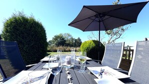 Tavolo da pranzo all'aperto apparecchiato con bicchieri trasparenti, piatti e posate, riparato da un ombrellone, con vista su un giardino e alberi.